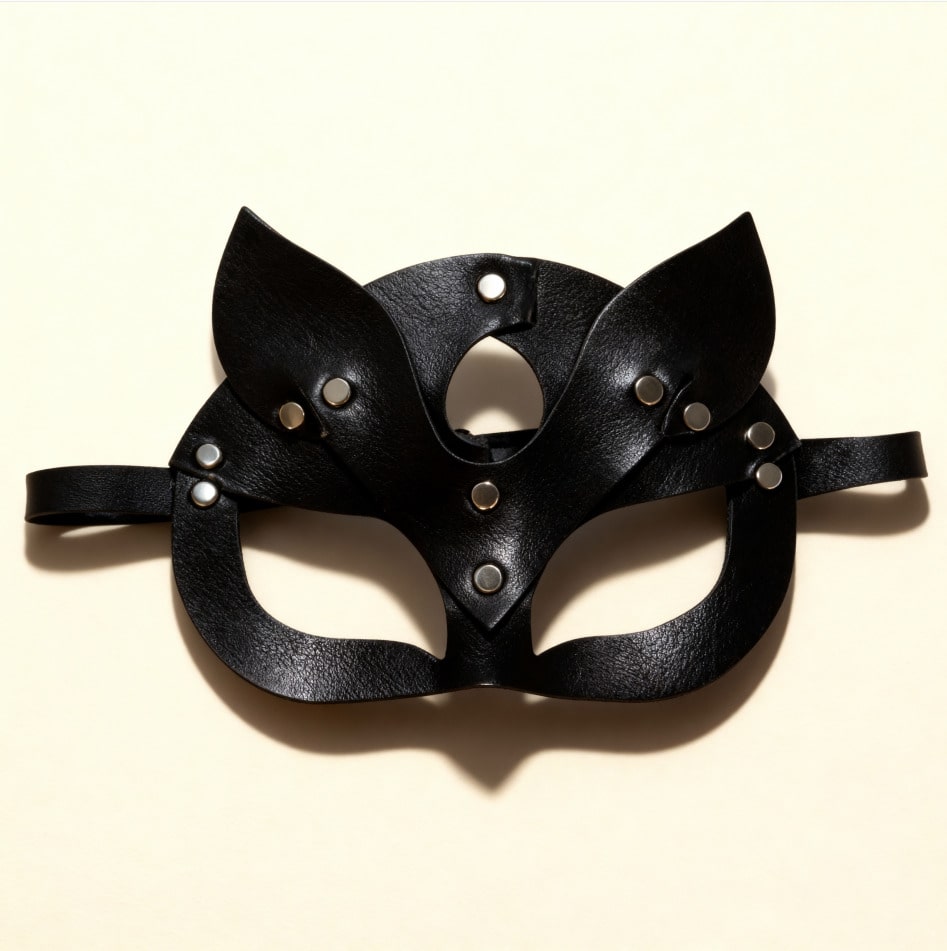 Masque en cuir noir style félin avec rivets métalliques, présenté à plat sur fond beige.